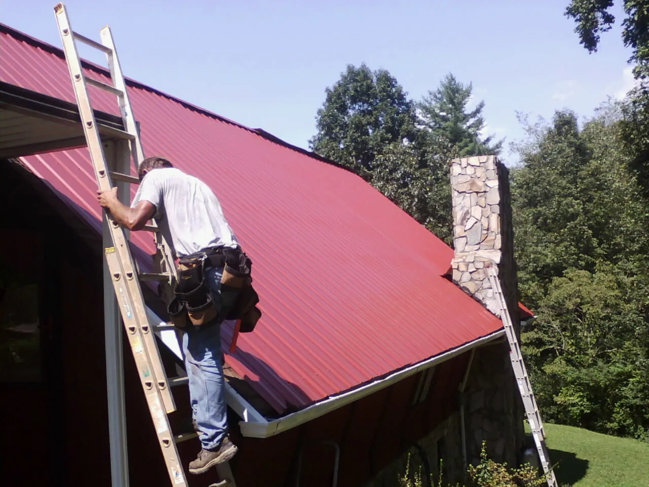 Roofer on a ladder beside a red metal roof and stone chimney — Soffit & Fascia Repair by Roofing Brothers in Kimballton